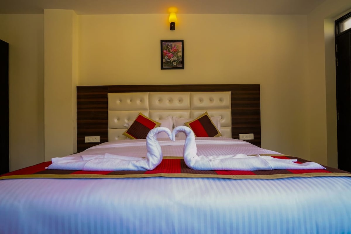 A comfortable sleeping area is presented, featuring a spacious bed adorned with an intricately folded towel arrangement resembling a heart. Complementary decorative pillows add a touch of color, while a framed floral artwork hangs above the headboard, enhancing the room's ambiance.
