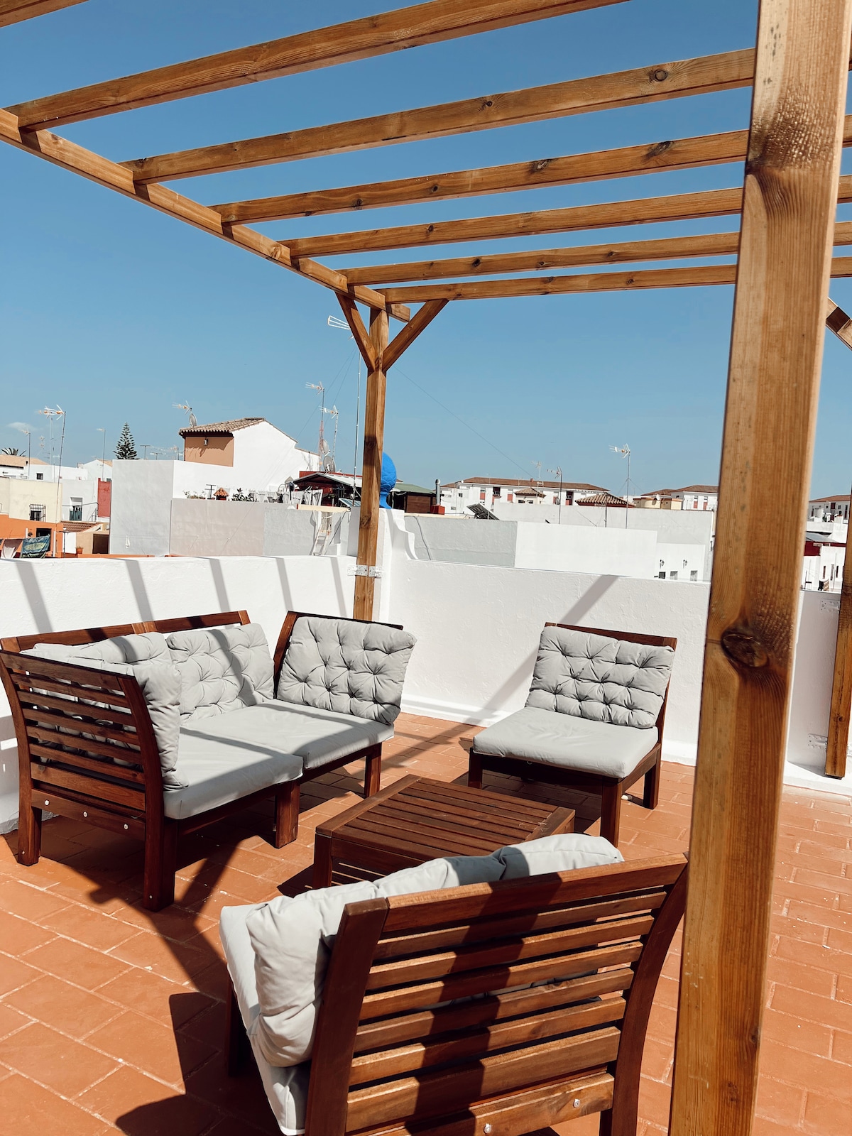 An outdoor terrace is featured, showcasing a wooden pergola overhead. The seating area includes four cushioned chairs arranged around a wooden coffee table. The bright blue sky serves as a backdrop, while the surrounding buildings are visible in the distance.