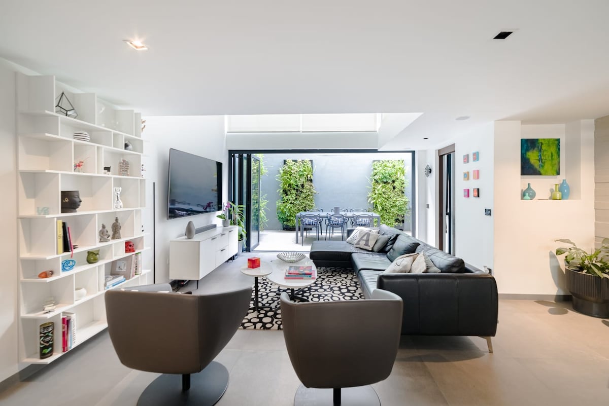 A modern living area features a sleek black sectional sofa facing a flat-screen television mounted on the wall. A stylish coffee table sits atop a patterned rug, while large glass doors open to a private outdoor space adorned with greenery and a dining set.