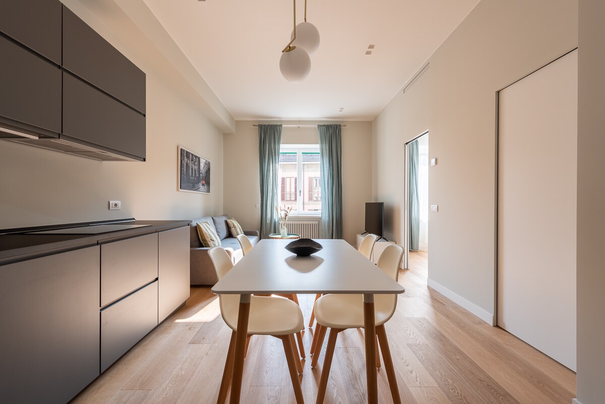 An open-plan living area features a modern kitchen with sleek cabinets and a table set for four. A comfortable sofa is positioned near large windows, which are dressed with soft curtains. Natural light enhances the warm tones of the wooden flooring.