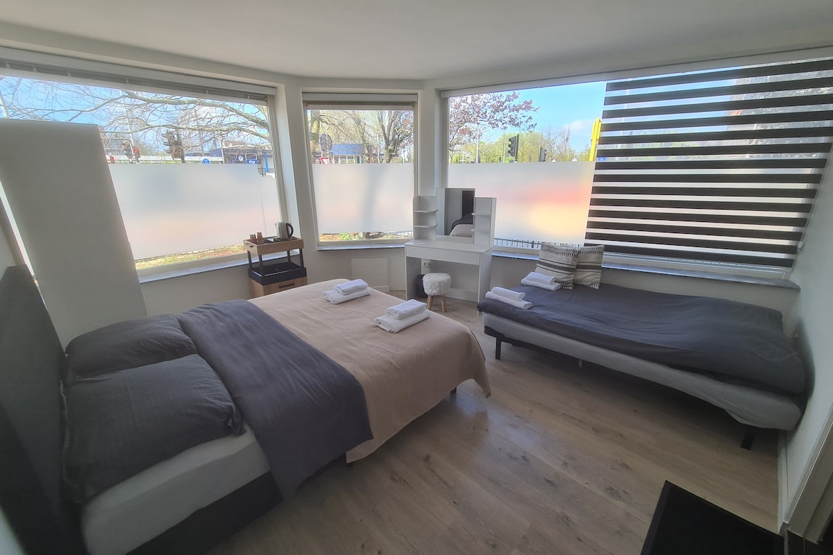 A cozy room features a spacious bed with a neutral-colored blanket and white linens. A comfortable sofa bed is positioned nearby. Large windows allow natural light to fill the space, and a small table with a kettle is also visible.