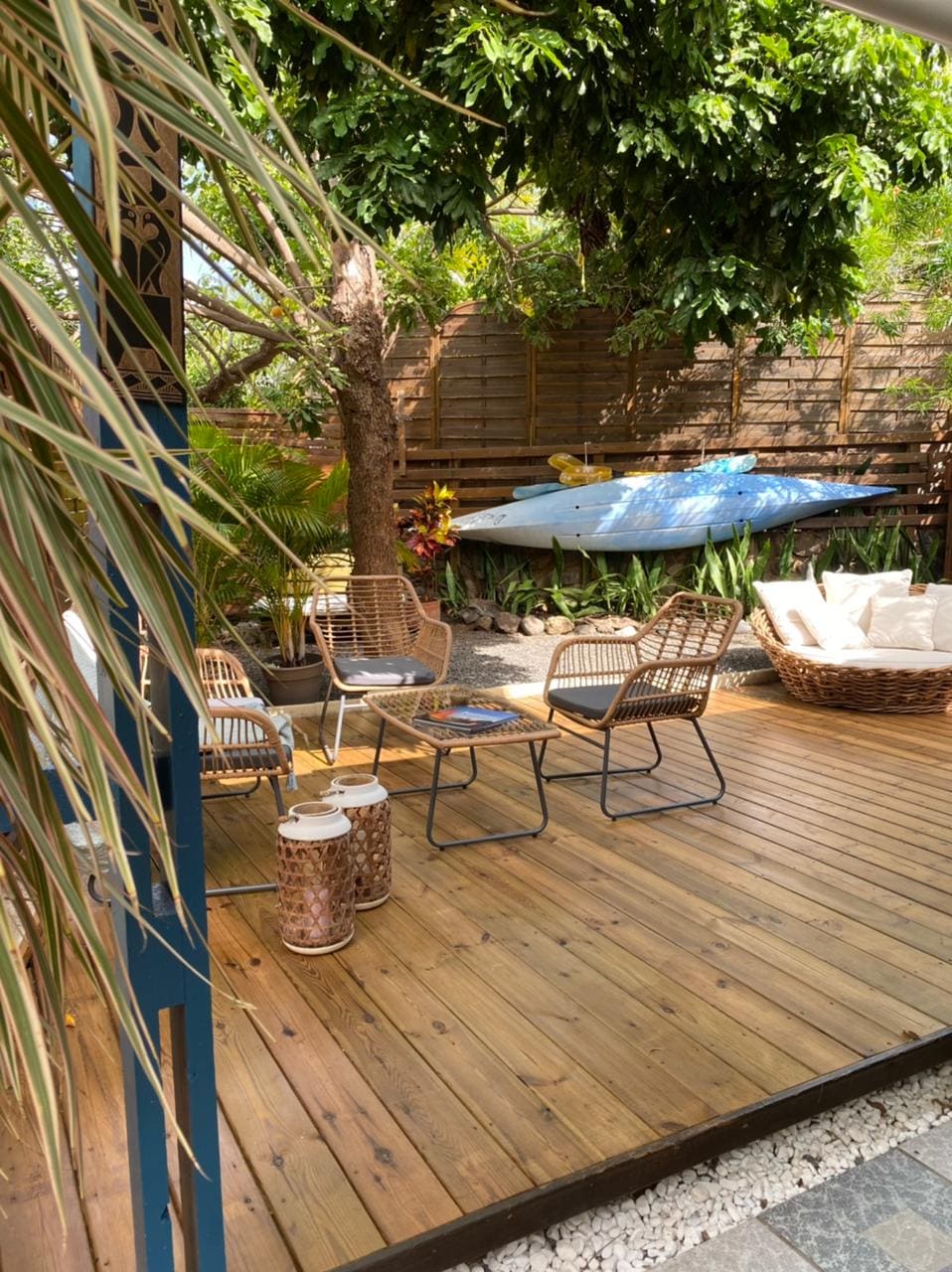 A wooden deck area is surrounded by lush greenery, featuring two woven chairs and a small table for relaxation. A kayak leans against the wall, while decorative lanterns and cushions provide a cozy touch. The space offers a serene atmosphere for outdoor enjoyment.