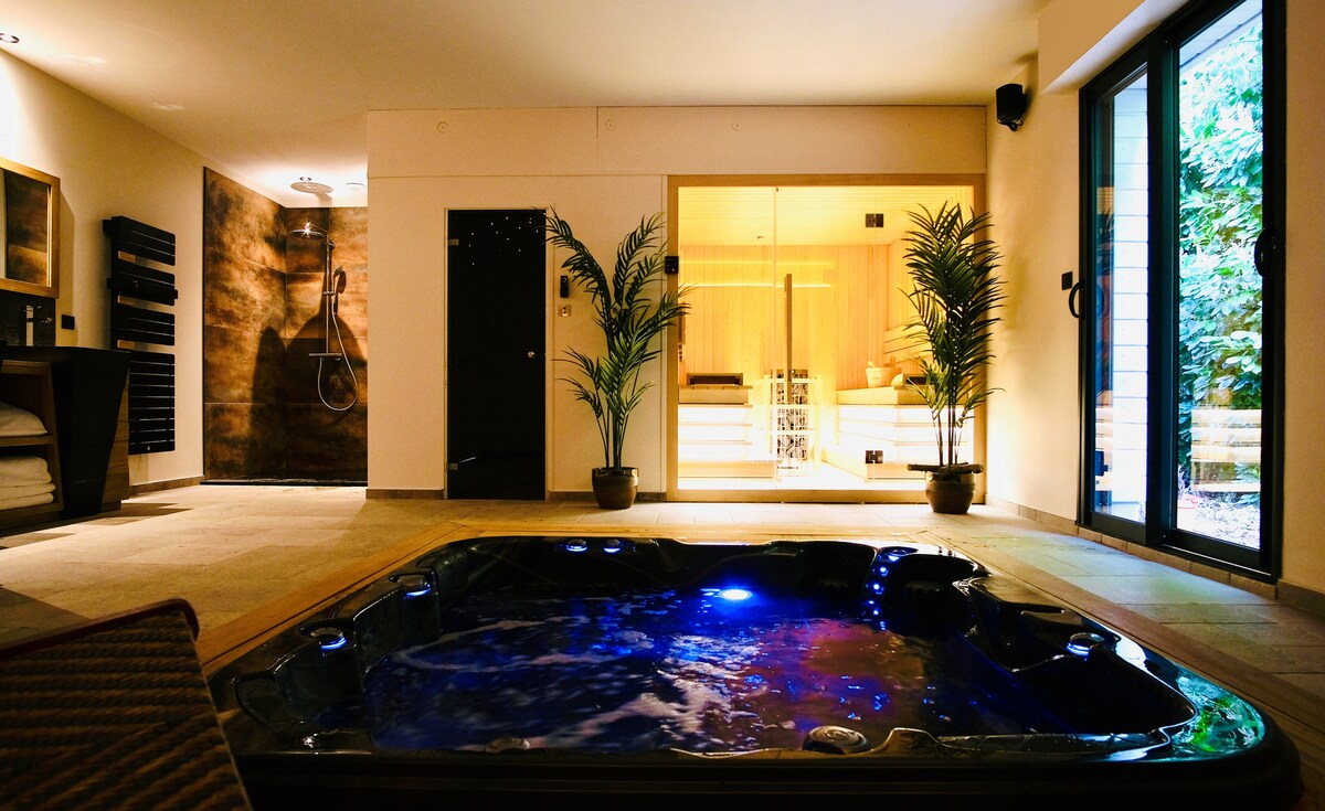 An indoor spa area features a jacuzzi with illuminated water, surrounded by potted plants. A spacious sauna is visible through glass doors, alongside a large shower area with a natural stone wall. Soft ambient lighting enhances the relaxing atmosphere.
