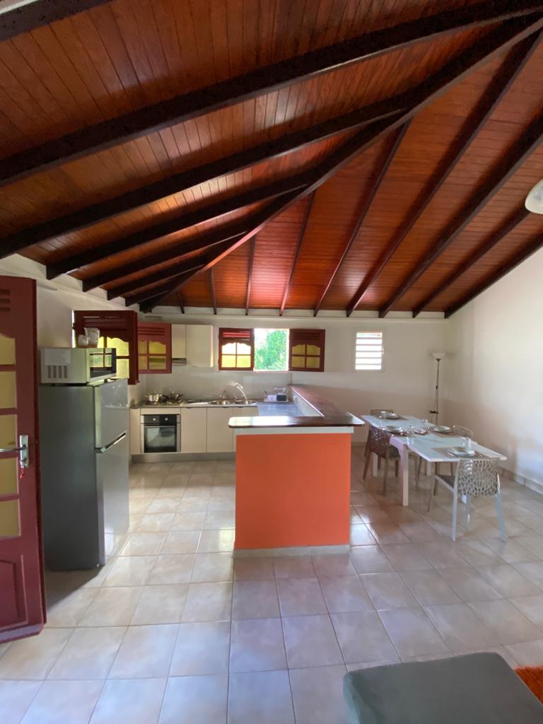 The kitchen area features a modern design with light-colored cabinets and appliances, complemented by a vibrant orange accent wall. A dining table with four chairs is provided, and large windows allow natural light to illuminate the space, highlighting the wood-beamed ceiling.