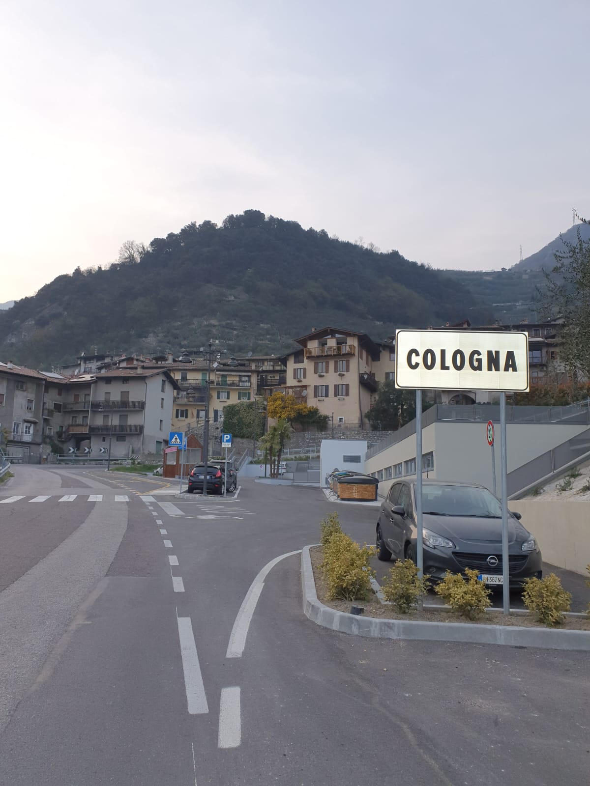 A view of Cologna is captured, featuring a road leading towards the village. In the foreground, a road sign displays 'COLOGNA.' Surrounding buildings showcase local architecture, while the mountainous backdrop provides a natural contrast to the urban setting.