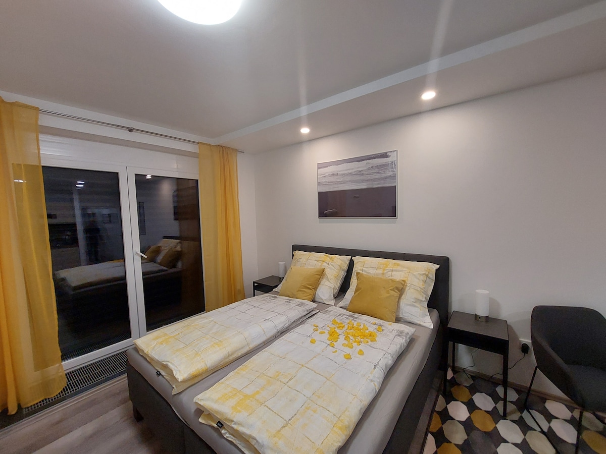 A well-lit bedroom is presented, featuring a spacious bed with neatly arranged linens. Yellow accents enhance the decor. A large glass door provides access to a balcony, while sheer curtains allow a gentle flow of natural light. A modern chair is visible in the corner.