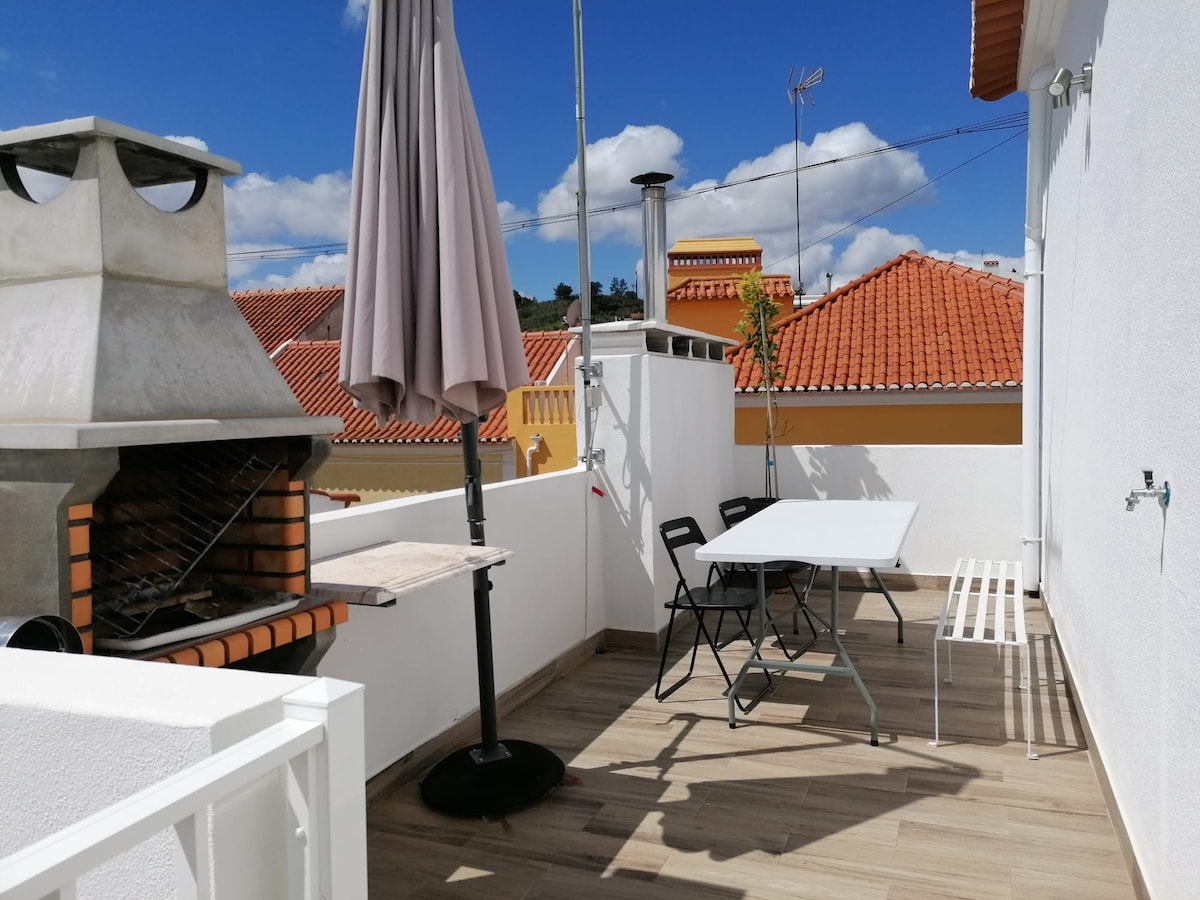 An outdoor terrace is shown with a barbecue grill and a round table surrounded by four chairs. A large umbrella provides shade. The view illustrates neighboring rooftops and a clear blue sky with scattered clouds.