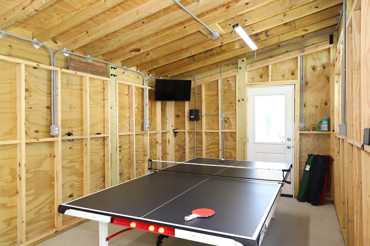 A game room is seen featuring a black ping pong table in the center. A television is mounted on the wall, and a door leads outside. The walls are constructed of light-colored plywood, and overhead lighting is present.