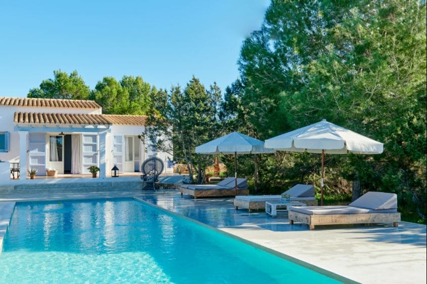 A tranquil outdoor area features a private pool surrounded by wooden loungers and shaded umbrellas. Lush greenery frames the space, with a white building visible in the background. Clear skies and sunlight enhance the serene ambiance of this Mediterranean retreat.