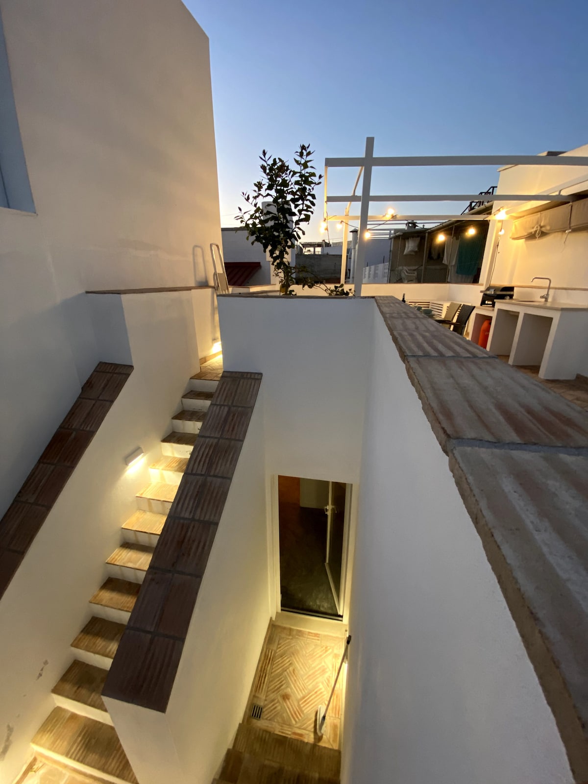 An exterior view reveals a set of illuminated stairs leading to a rooftop terrace. The staircase is flanked by white walls and a potted plant, while outdoor seating and cooking facilities can be seen on the terrace above, all under a dusk sky.