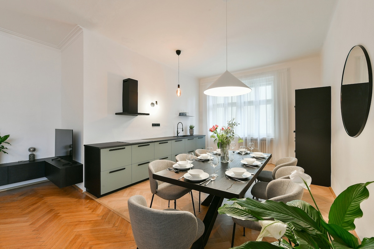 A modern dining area showcases a large black table surrounded by comfortable gray chairs. A stylish pendant light hangs overhead, and a mirror is mounted on the wall. The room features light wood flooring and large windows that allow natural light to fill the space.