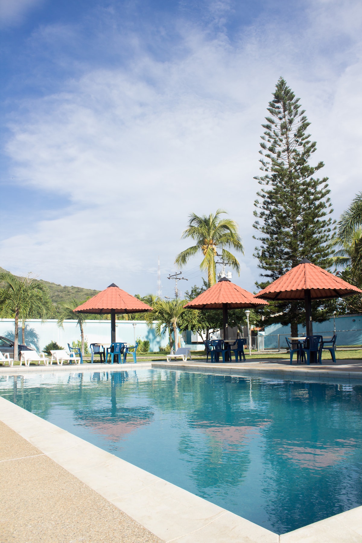 A well-maintained outdoor pool is surrounded by lush greenery and palm trees. Two canopy-covered seating areas accommodate several tables and chairs, providing shaded spots for relaxation. The pool's clear blue water reflects the sky, enhancing the tranquil atmosphere of the garden area.