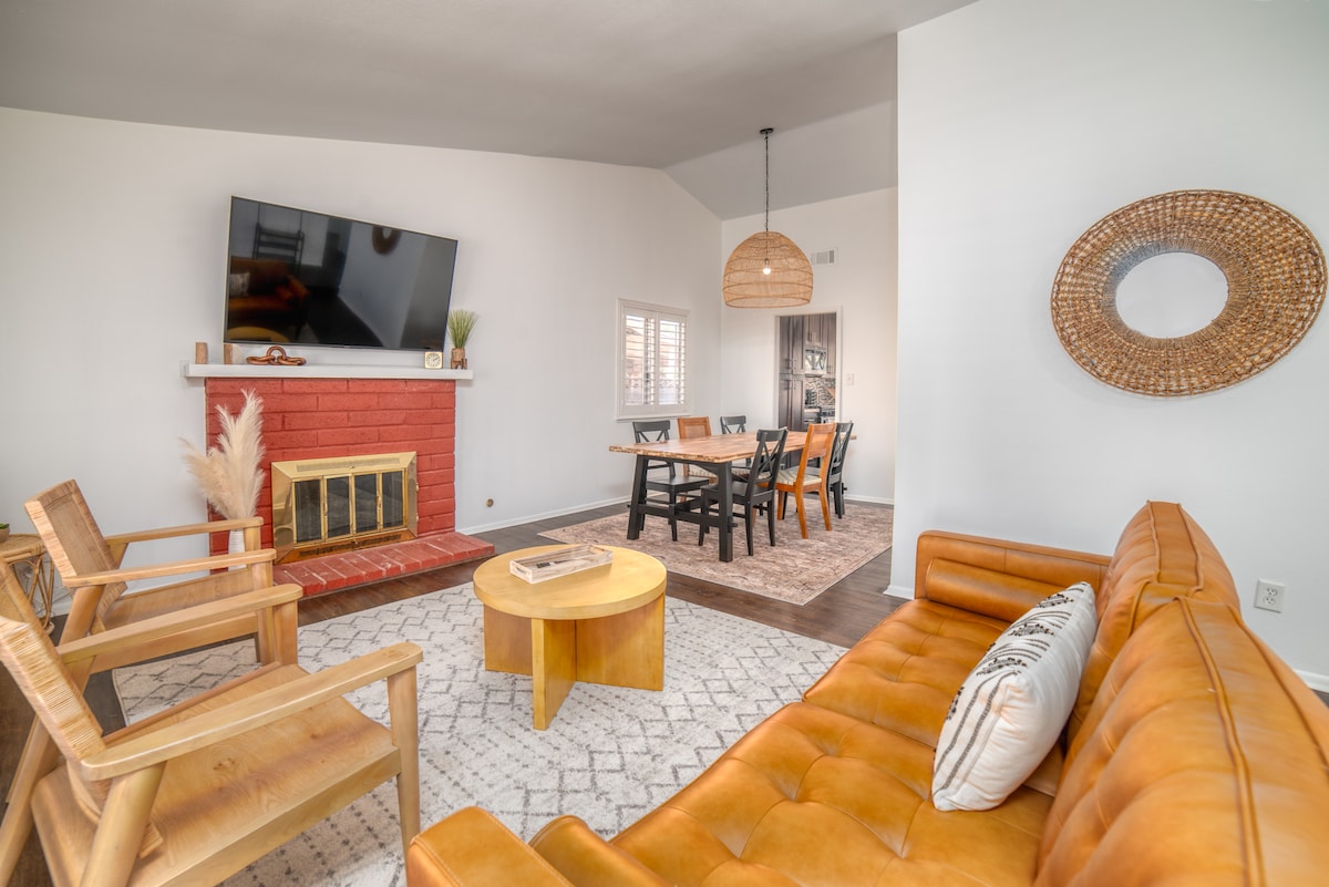 An inviting living space features a warm-toned leather sofa complemented by wooden armchairs. A round coffee table sits atop a textured rug. A red-brick fireplace serves as a focal point beneath a mounted flat-screen TV, while natural light filters in through nearby windows.