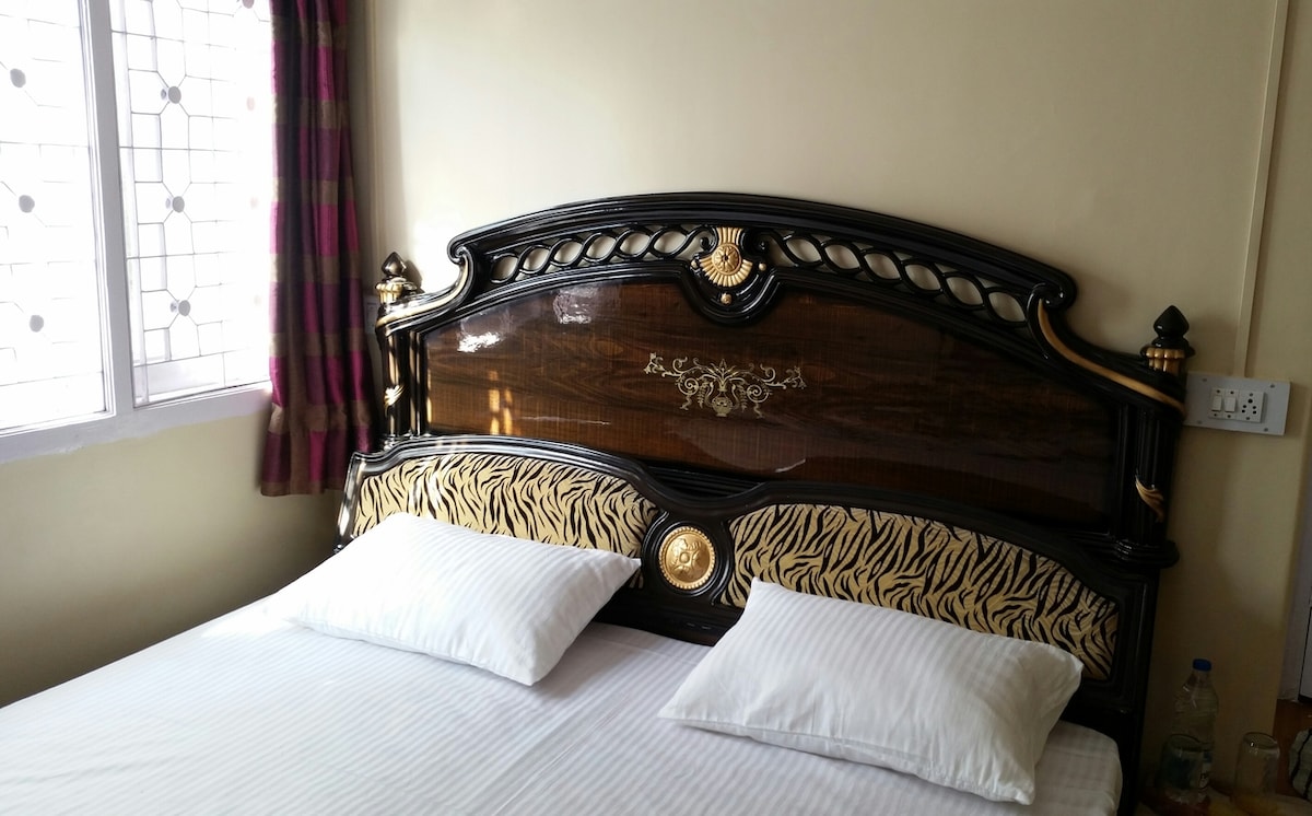 A stylish headboard featuring intricate designs is paired with clean white linens on the bed. Two pillows are neatly arranged at the top of the mattress. Natural light enters through a window adorned with patterned curtains, enhancing the room's bright and inviting atmosphere.