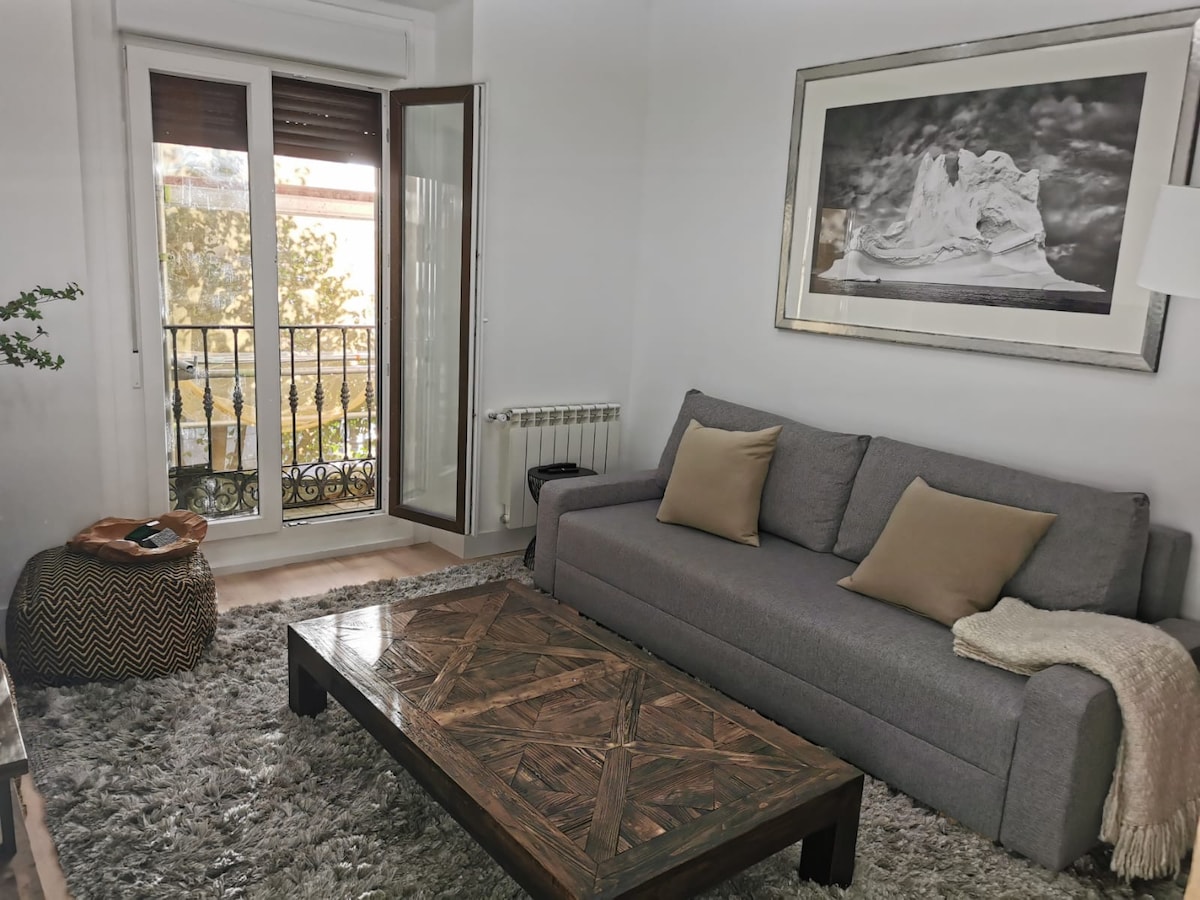 A comfortable living area features a gray sofa with soft cushions and a textured throw draped over one side. A wooden coffee table with a patterned surface sits on a plush rug. Large windows, partially open, frame a view of the outside balcony.