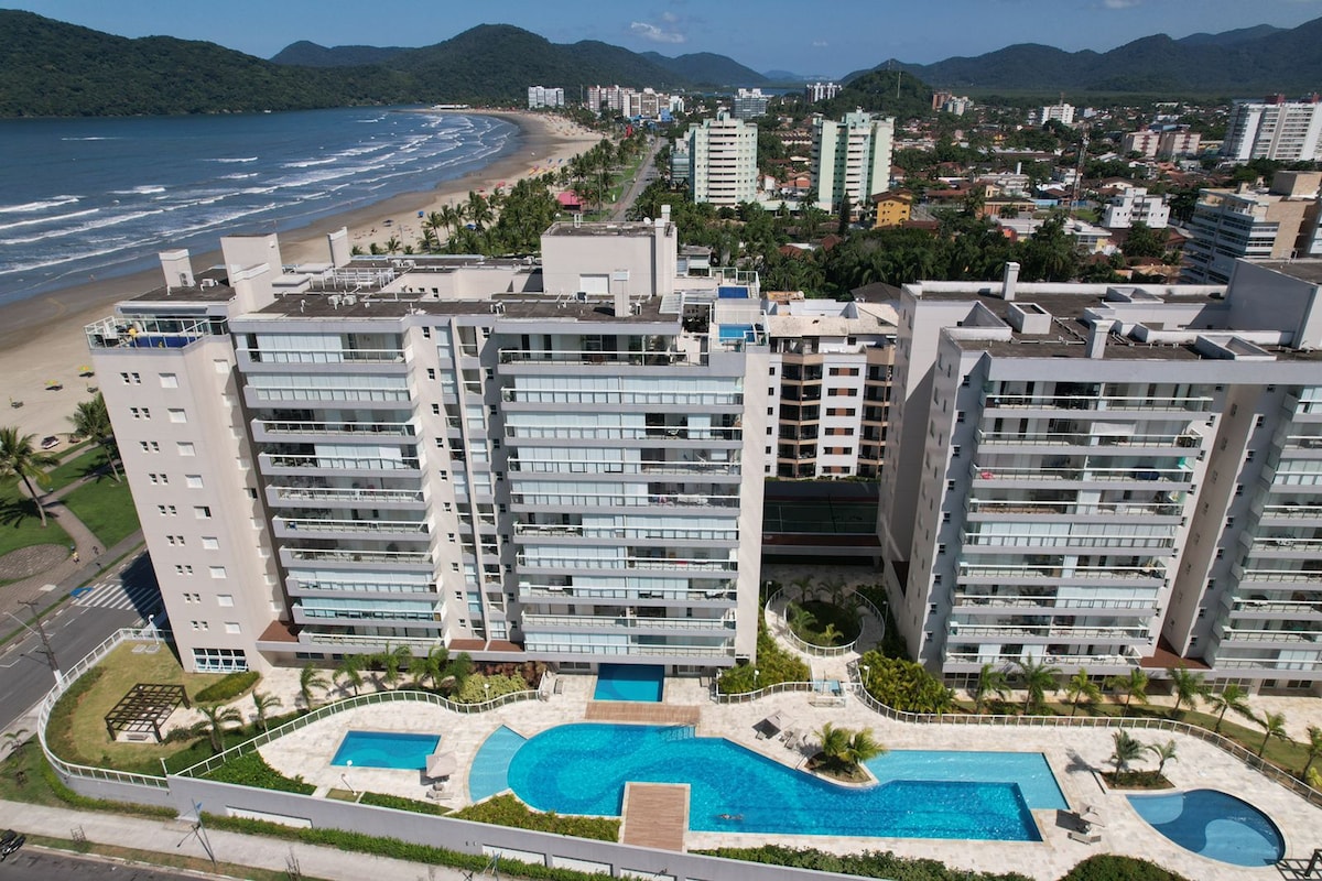 An aerial view of a modern condominium complex reveals a spacious pool area surrounded by landscaped green spaces. The coastline and beach are visible in the background, along with high-rise buildings and lush mountains in the distance, creating a picturesque tropical setting.
