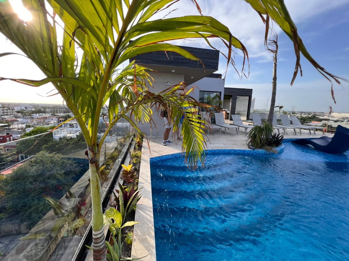 A modern rooftop pool area is set against a vibrant city backdrop. Lush palm plants frame the space, while a sparkling blue pool invites relaxation. Comfortable lounge chairs are arranged around the pool, allowing for leisurely soaking up the sun.