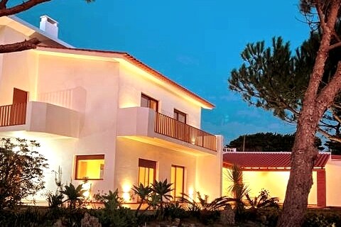 The exterior of a white two-story house is illuminated at dusk, highlighting its sleek architecture. Balconies are visible on the upper level, and lush greenery surrounds the property, adding to the calm ambiance of the setting.