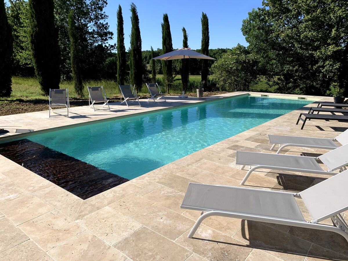 A serene salt-water pool is framed by a spacious terrace featuring several lounge chairs arranged for relaxation. Lush greenery and tall cypress trees provide a natural backdrop, while a shaded umbrella offers respite from the sun.