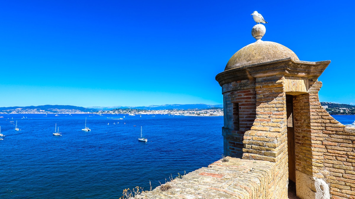 A coastal view is captured from a historic stone structure, featuring a round turret topped with a bird. The calm blue waters stretch towards the horizon, dotted with sailboats, while the scenic coastline of Cannes is visible in the background under a clear sky.