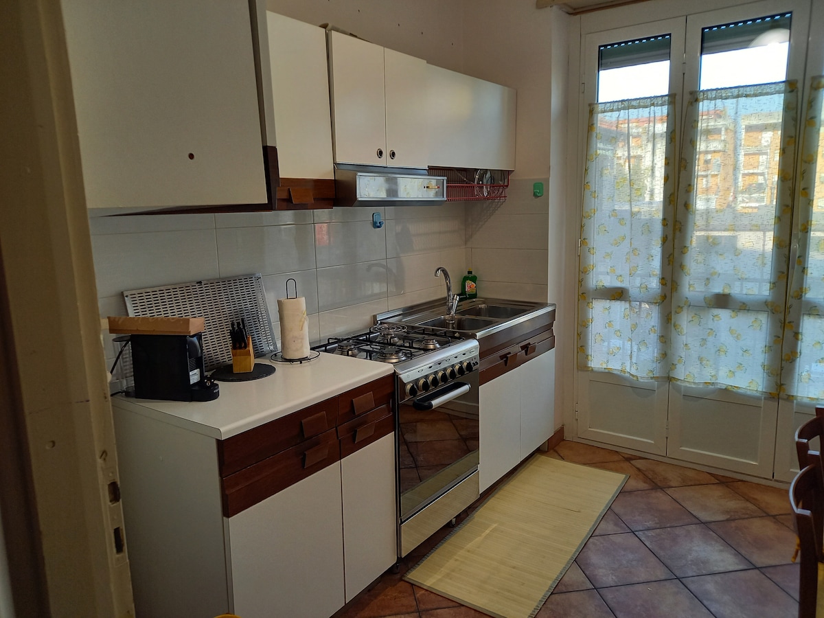 A functional kitchen features white cabinets with wooden accents and a gas stove. Natural light enters through a window adorned with a patterned curtain. A counter is equipped with essential kitchen tools, creating a practical cooking space.
