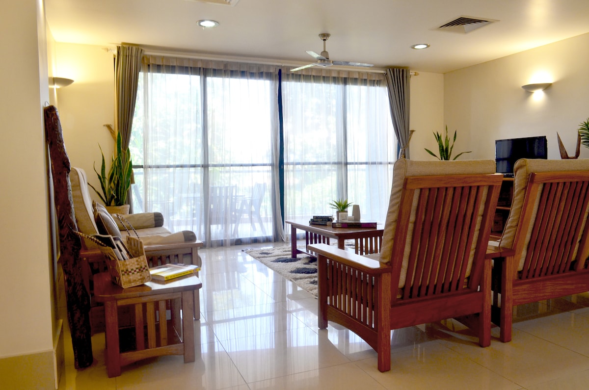 A spacious living area is filled with natural light streaming through large glass doors. Wooden seating arrangements are complemented by potted plants. A coffee table sits centrally on a patterned rug, with a view of the outdoors visible through the sheer curtains.