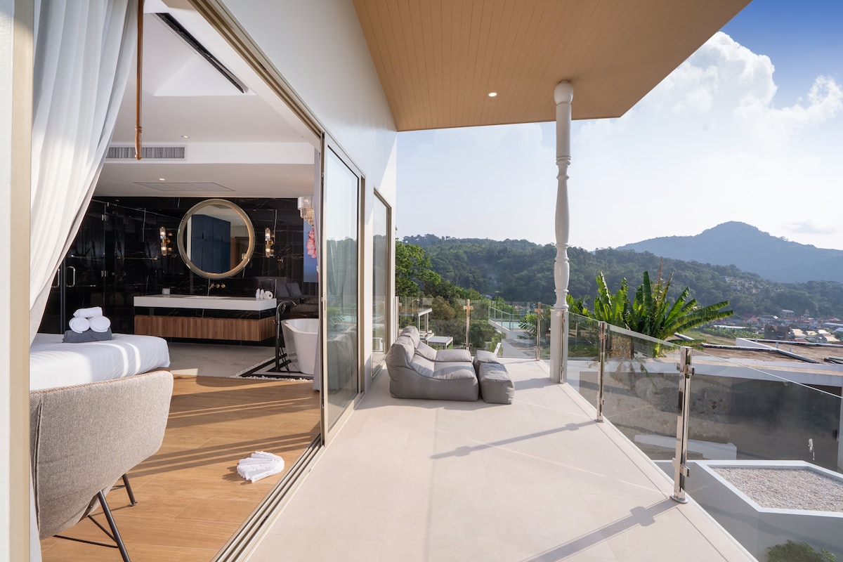 A spacious balcony is featured, offering views of the surrounding hills. The balcony includes comfortable seating and is accessed directly from a bedroom. Sliding glass doors lead to an elegant interior space that showcases modern design elements and a well-appointed ensuite bathroom.