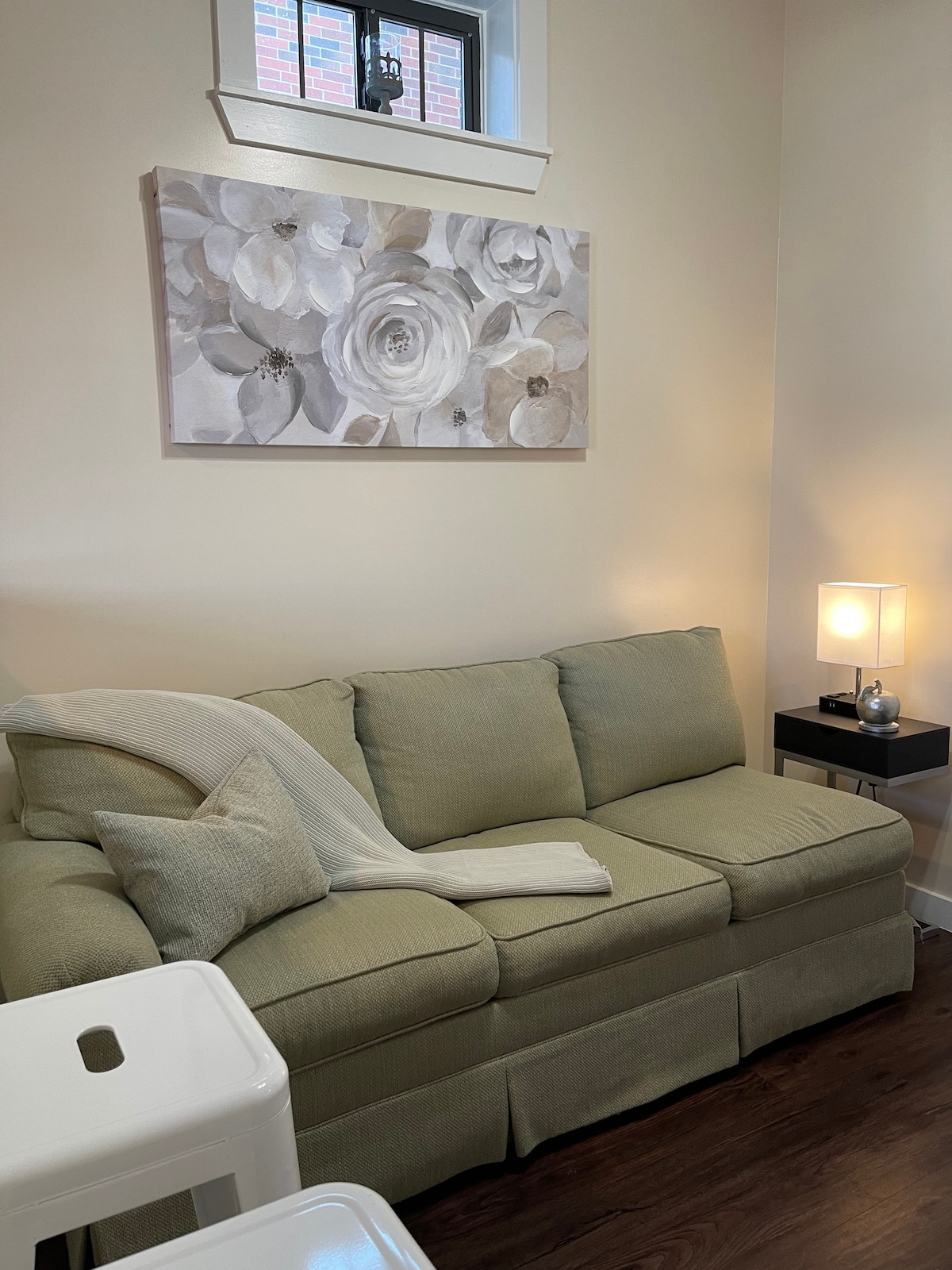 A soft green sofa is positioned in the living area, complemented by a light throw and decorative pillows. Above the sofa, a neutral-toned floral artwork adds a touch of elegance. A sleek side table with a lamp provides subtle lighting, enhancing the room's cozy feel.