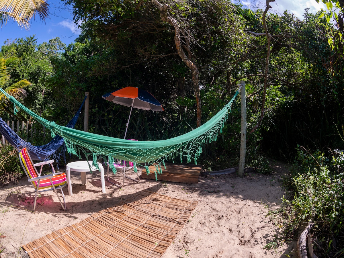 A hammock in vibrant green fabric is suspended between two posts. Nearby, a small white table is accompanied by colorful chairs. A beach umbrella provides shade, while a sandy area is partially covered by a bamboo mat. Lush greenery surrounds the space, contributing to a serene setting.