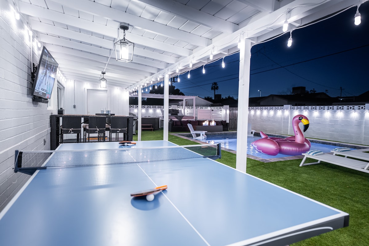An outdoor entertainment area is illuminated by string lights, featuring a ping pong table ready for play. A large television is mounted on the wall, and comfortable seating is arranged nearby. A pink inflatable flamingo floats in the heated pool, surrounded by lush green turf.