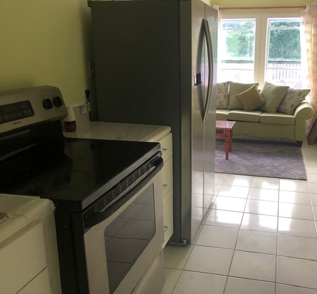 A bright kitchen area showcases modern appliances, including a silver refrigerator and a black stove. The adjacent living space features a cozy couch adorned with cushions, with natural light filtering through large windows, enhancing the bright and airy atmosphere.