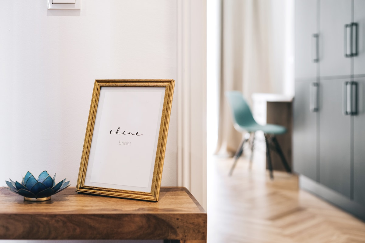 A framed print with the word 'shine' in elegant script is displayed on a wooden table. Behind it, a muted area of the room shows a chair and cabinetry, while natural light filters through soft curtains.