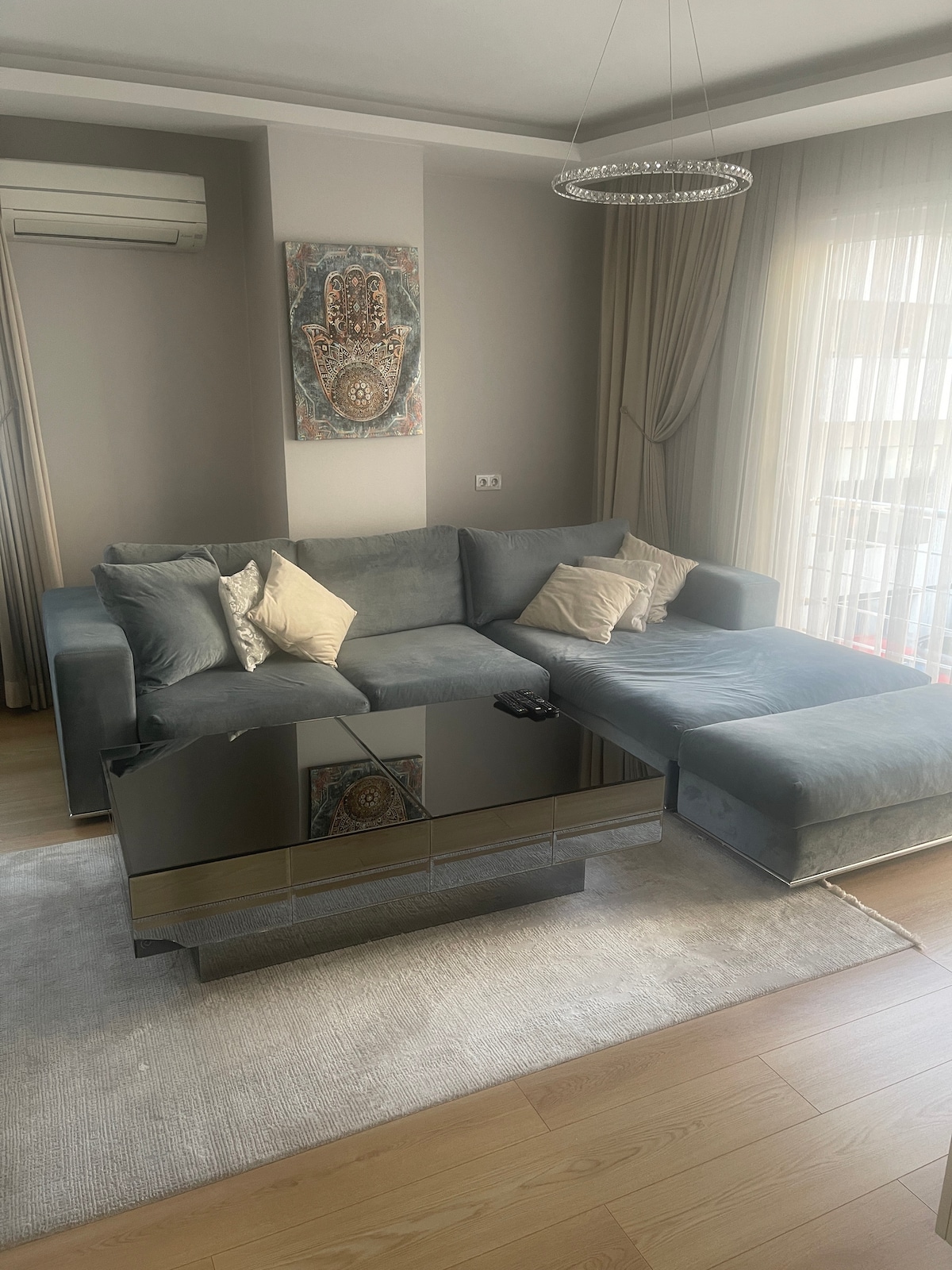 A comfortable lounge area features a grey sectional sofa adorned with two accent pillows. A mirrored coffee table sits at the center, reflecting natural light from the adjacent windows. Light-coloured curtains gently frame the windows, complementing the warm wooden flooring.