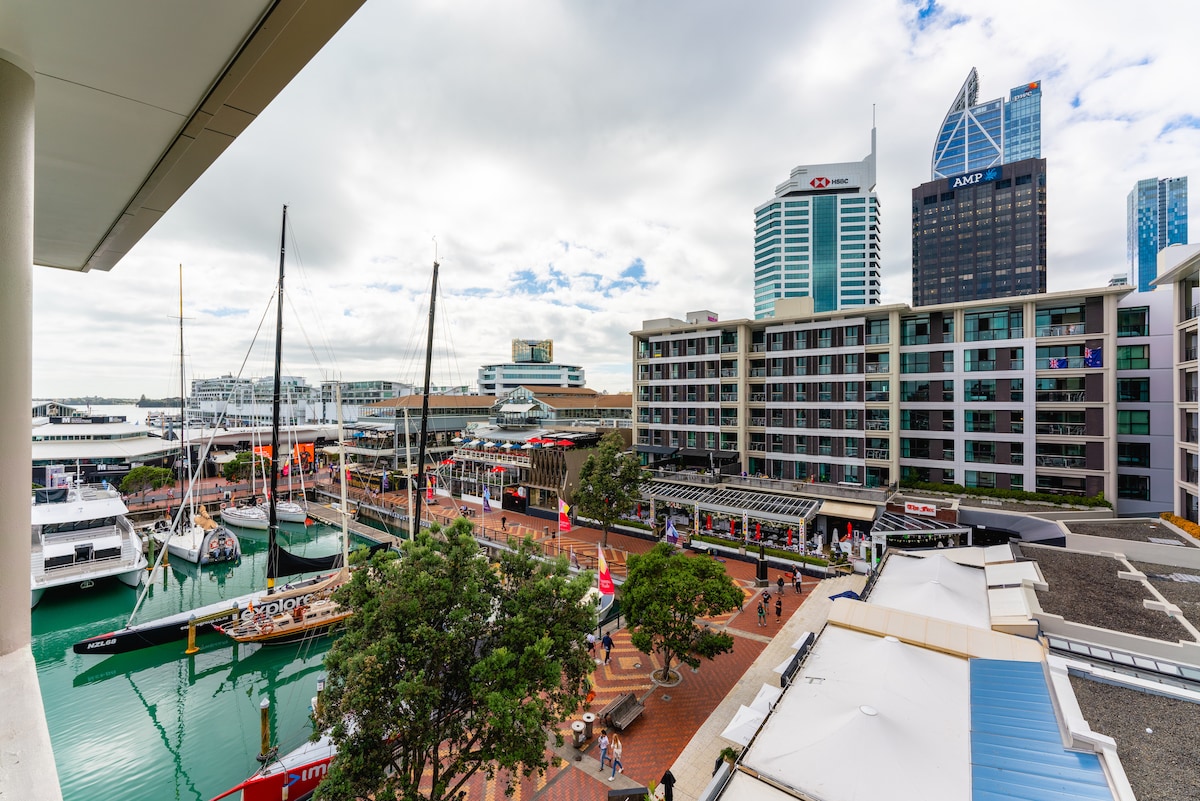 A panoramic view from a balcony reveals the bustling viaduct with luxury yachts docked in the marina. Modern buildings with an array of shops line the waterfront, while pedestrians enjoy the vibrant atmosphere below. The scene is framed by overcast skies above.