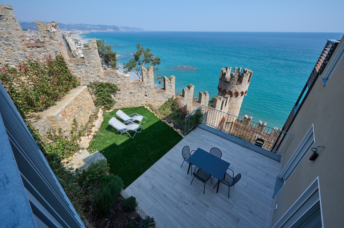 A spacious outdoor terrace overlooks the sea, featuring a dining table set and lounge chairs positioned on green grass. The scene is framed by ancient castle walls, enhancing the coastal view and natural surroundings.