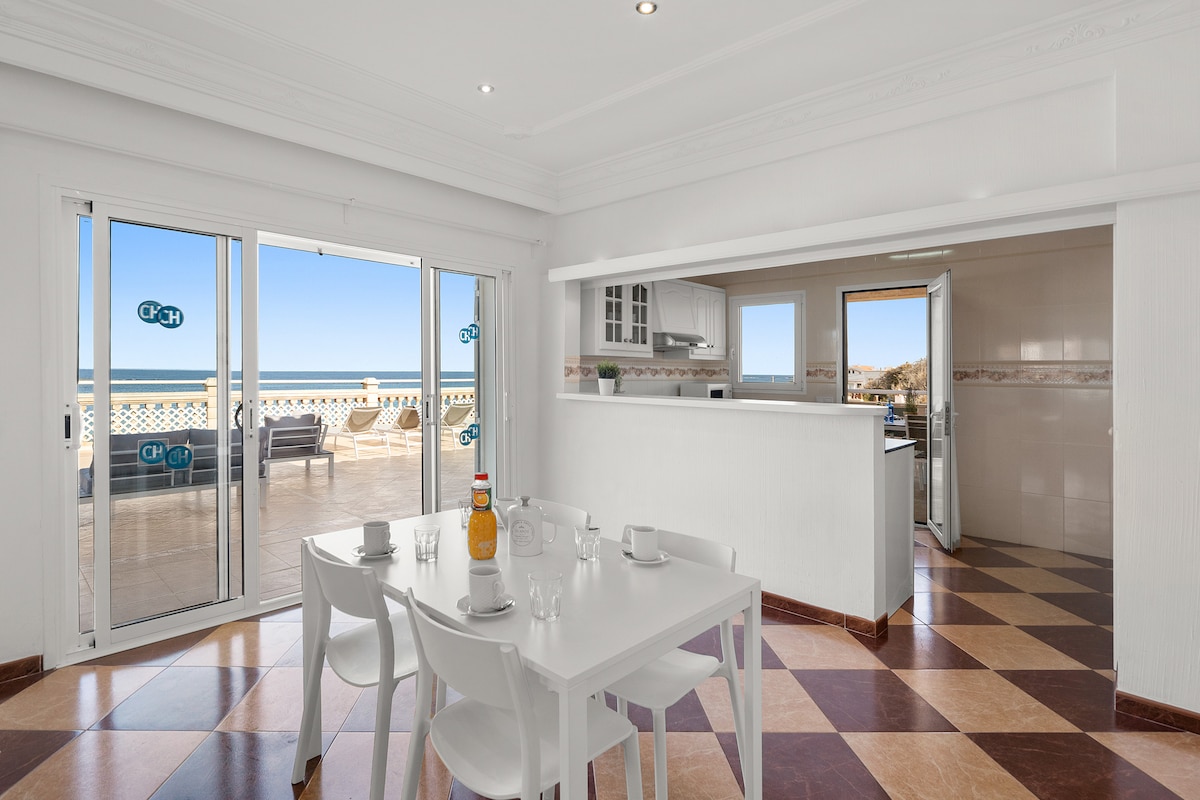 A bright dining area features a white table set for four, with a bottle of drink in the center. Large sliding doors provide access to the terrace and showcase ocean views. The kitchen is visible in the background, with modern cabinetry and appliances.