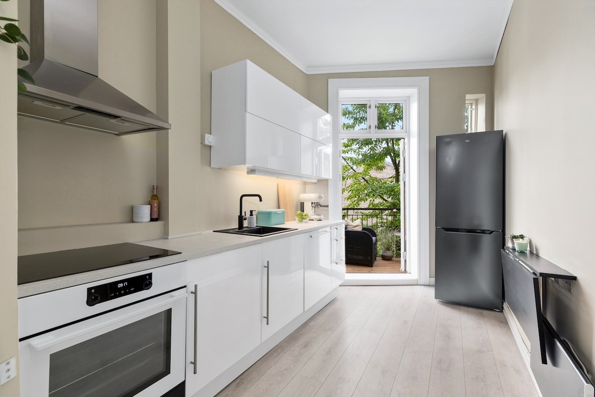 A modern kitchen features sleek, white cabinetry and stainless steel appliances. Natural light enters through a large window that leads to a small balcony. A refrigerator stands against the wall, while the countertop offers ample workspace for meal preparation.