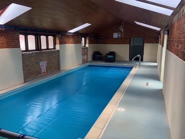 An indoor swimming pool is illuminated by natural light from skylights. The pool is covered, and a smooth, light-colored surface surrounds it. Comfortable seating areas are positioned beside the pool, creating a relaxing environment.