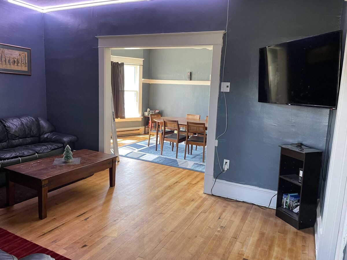 A living area features dark hardwood flooring and a leather sofa, complemented by a wooden coffee table. A doorway leads to a dining area with a blue rug, and a television is mounted on the wall, providing modern convenience in a classic setting.