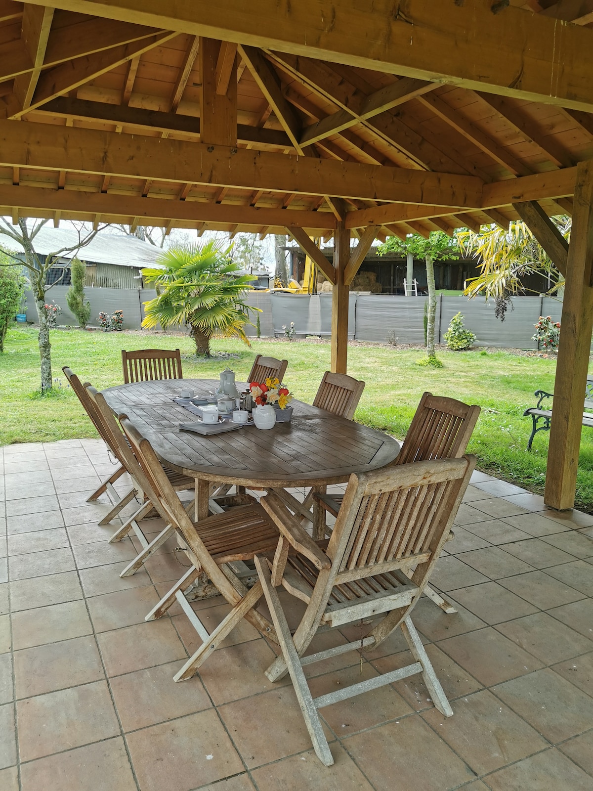 A wooden outdoor dining table with matching chairs is situated beneath a covered gazebo. Surrounding greenery, including palm trees and flower beds, adds a natural setting. The tiled floor provides a stable surface for meals, enhancing outdoor dining opportunities in the scenic environment.