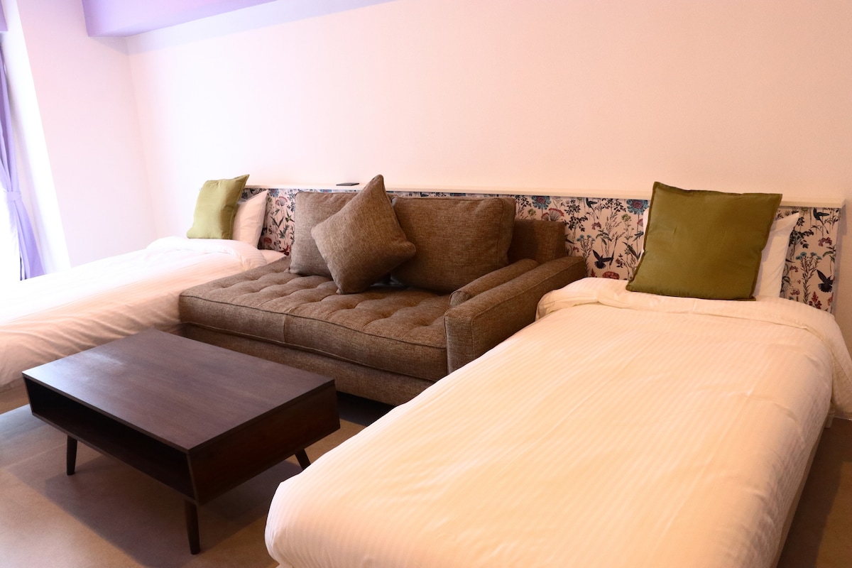 A room is presented with two spacious beds and a stylish sofa. The sofa features decorative cushions and floral upholstery, complementing the room's bright and airy atmosphere. A wooden coffee table is positioned in front of the seating area, adding functionality to the space.