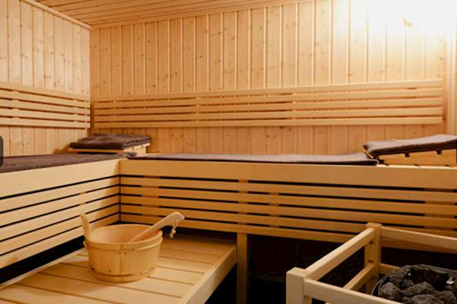 The sauna features wooden benches arranged around a simple stone heater. A wooden bucket sits on one corner, with soft, dark towels neatly placed on the benches. The warm wood paneling contributes to a calming atmosphere.