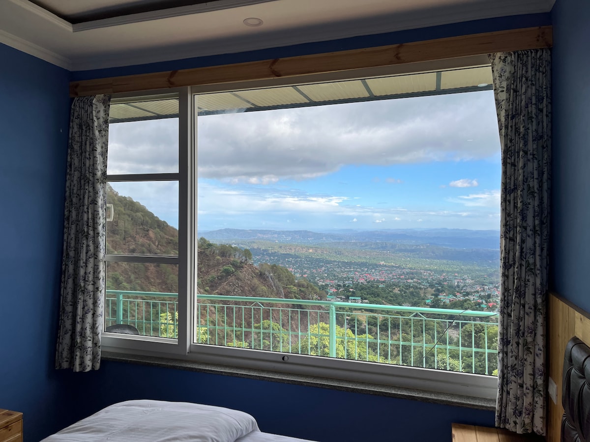 A large window frames a breathtaking view of the hills and valley beyond. The surrounding blue walls and patterned curtains complement the serene landscape, with lush greenery and distant mountains visible under a partly cloudy sky.