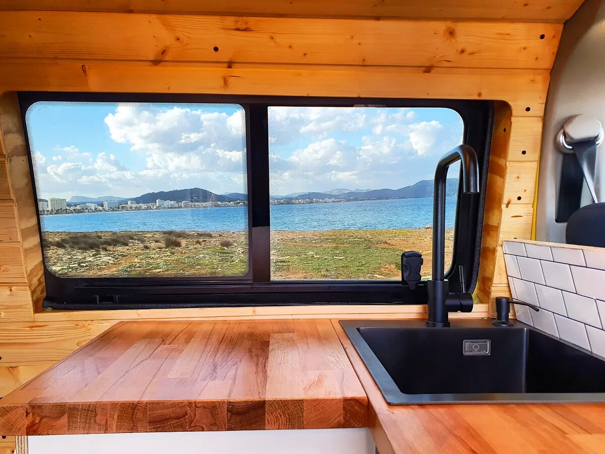 A view of the exterior landscape is framed by a large window in the camper van, showcasing a serene waterscape with distant mountains under a partly cloudy sky. The wooden countertop and modern sink are prominently featured in the foreground.