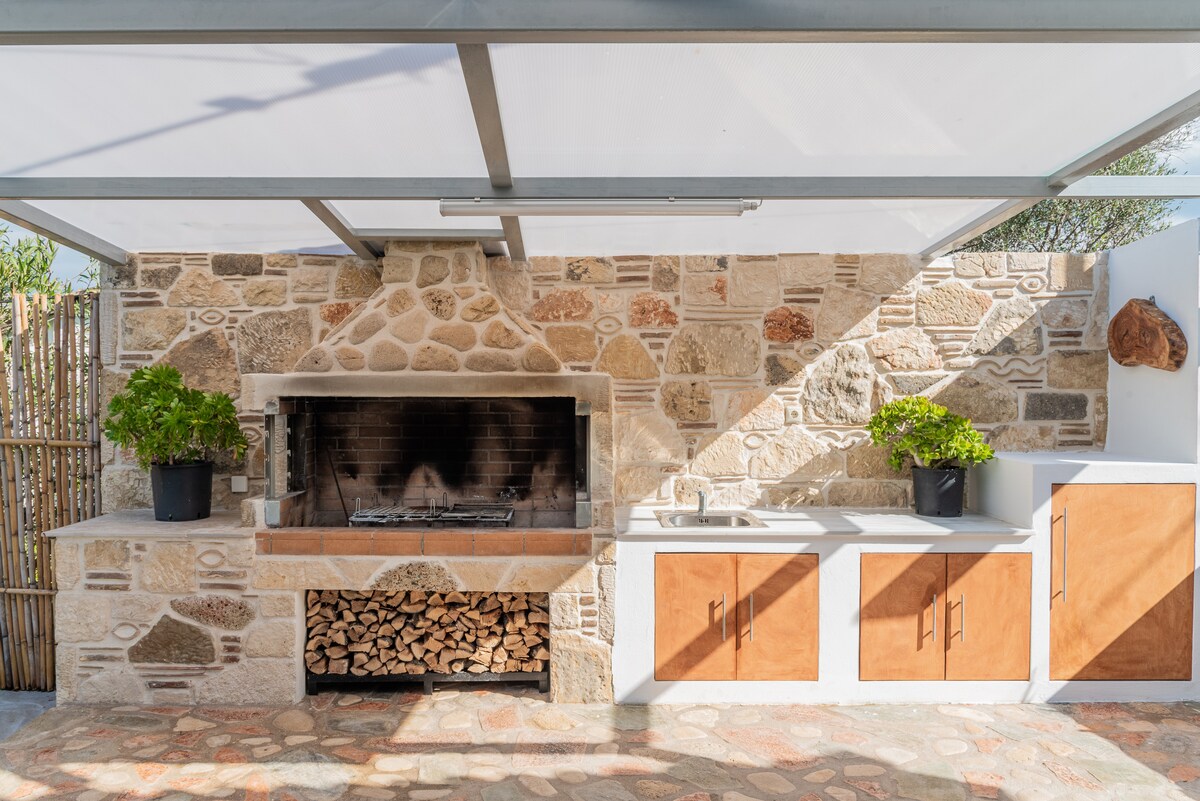 An outdoor barbecue area is equipped with a stone grill and a wood storage compartment. The kitchen counter features a sink, complemented by potted plants on either side. Natural stone walls surround the space, providing a rustic charm.