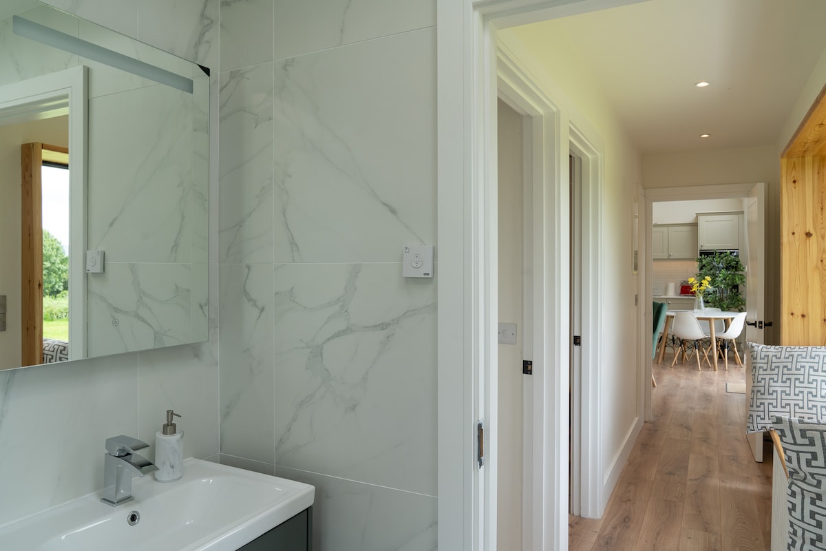 A modern bathroom features marble-patterned walls and a sleek vanity with a mirror above. A doorway in the background leads to an inviting hallway, with natural light illuminating the space and hints of greenery visible outside through the windows.