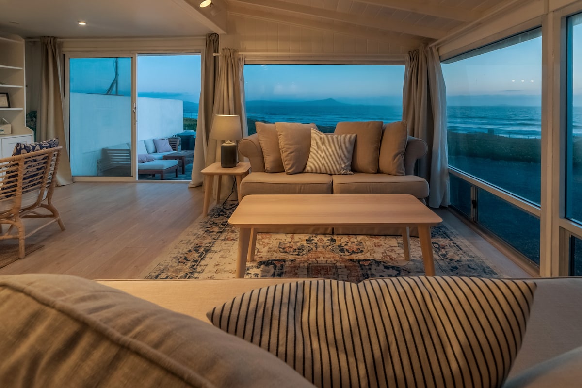 An open living space features a comfortable beige sofa with patterned cushions, facing a light wood coffee table. Large windows offer expansive views of the beach and bay, with soft, natural light illuminating the airy interior. A cozy chair is positioned nearby.