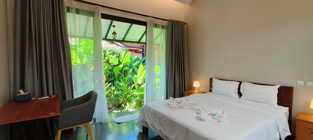 A spacious bedroom features a large bed adorned with white linens and decorative towel arrangements. Soft lighting is provided by two bedside lamps. Floor-to-ceiling glass doors open to a lush garden, while dark curtains frame the view. A wooden desk and chair are positioned near the entrance.