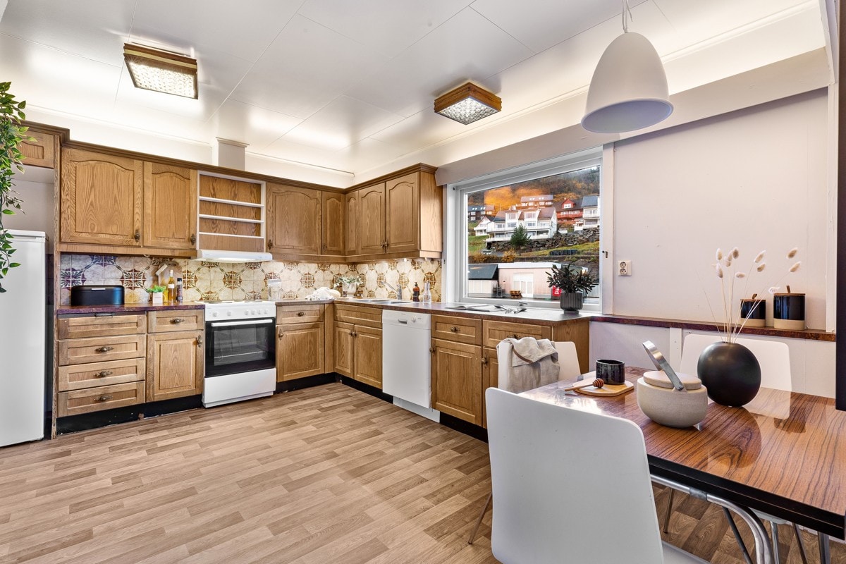 The kitchen features wooden cabinetry and a tiled backsplash, with modern appliances including a stove, dishwasher, and refrigerator. A dining table with a decorative centerpiece is present, alongside a window showcasing views of the surroundings, allowing natural light to fill the space.