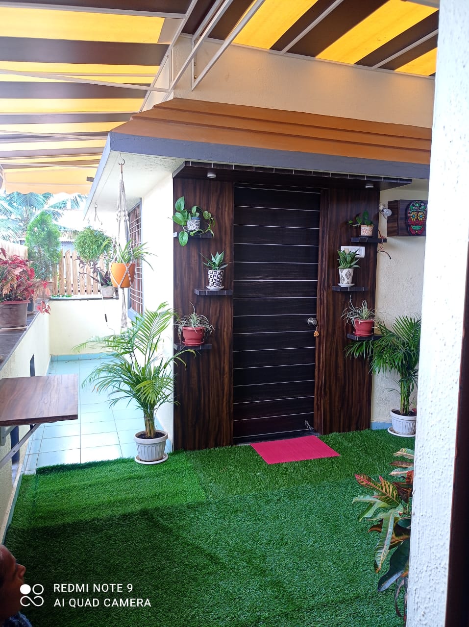 An entrance area is framed by vibrant greenery and potted plants. The doorway features a dark wood panel with a small red mat in front. Overhead, a striped awning provides shelter, while artificial grass offers a natural touch to the space.